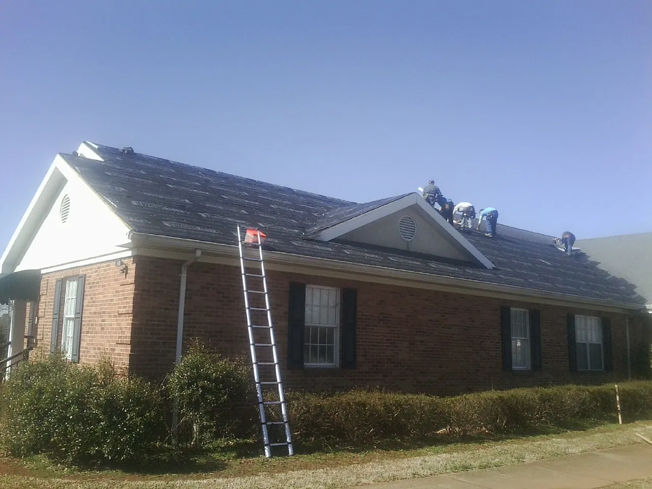 Crew tearing shingles off a brick ranch roof with a ladder set — Emergency Roof Repair work by our crew in Little Mountain