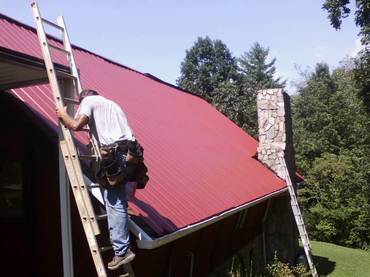 Roofer on a ladder beside a red metal roof and stone chimney — Soffit & Fascia Repair by Roofing Brothers in Little Mountain